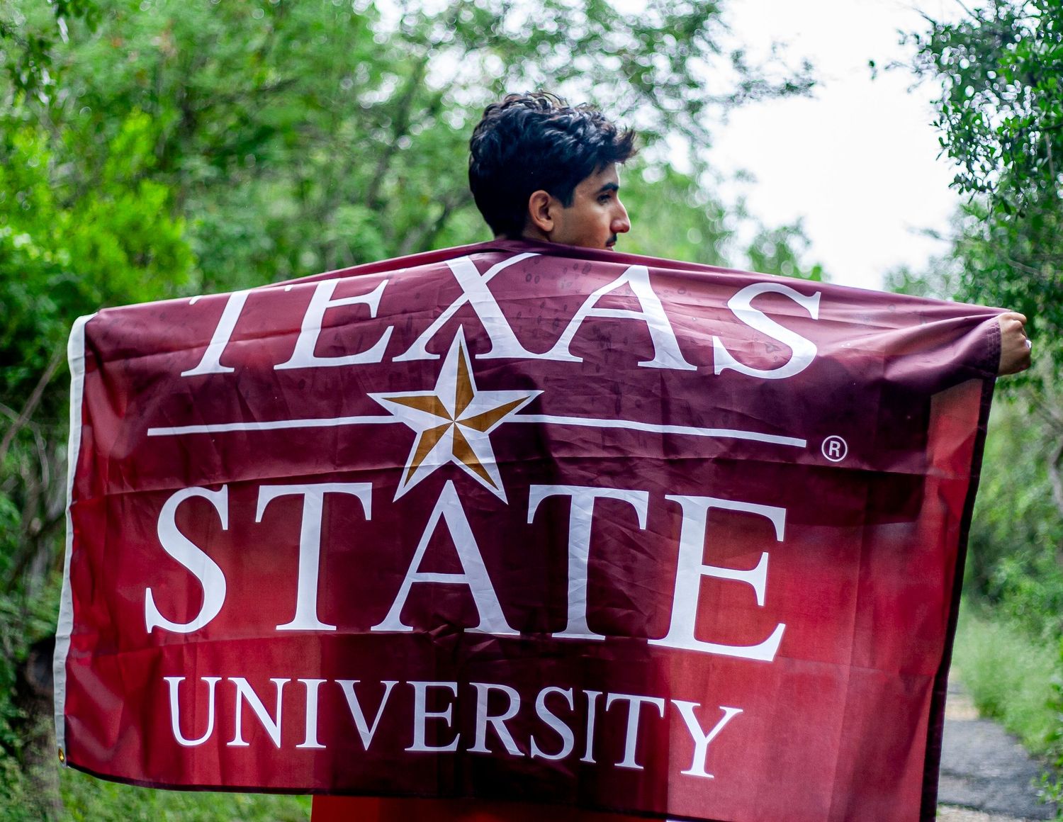 A maroon Texas State University flag with star logo displayed in a wooded outdoor setting.