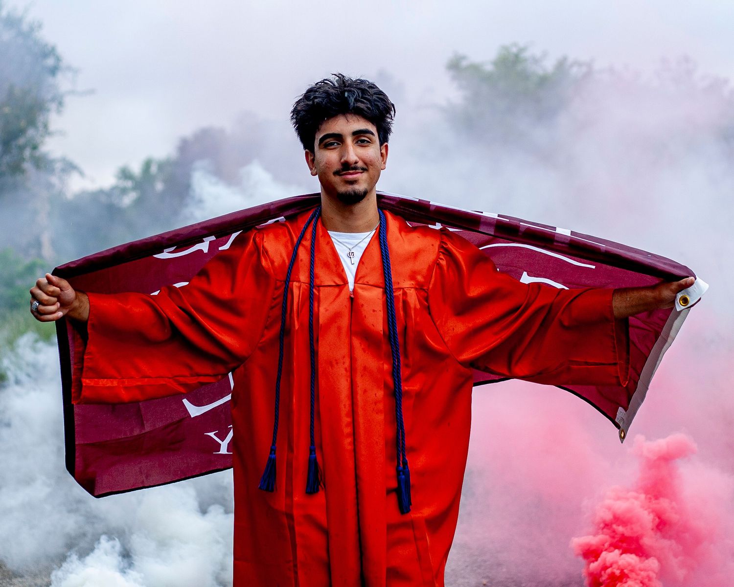 A graduate in red regalia poses with smoke effects in the background.