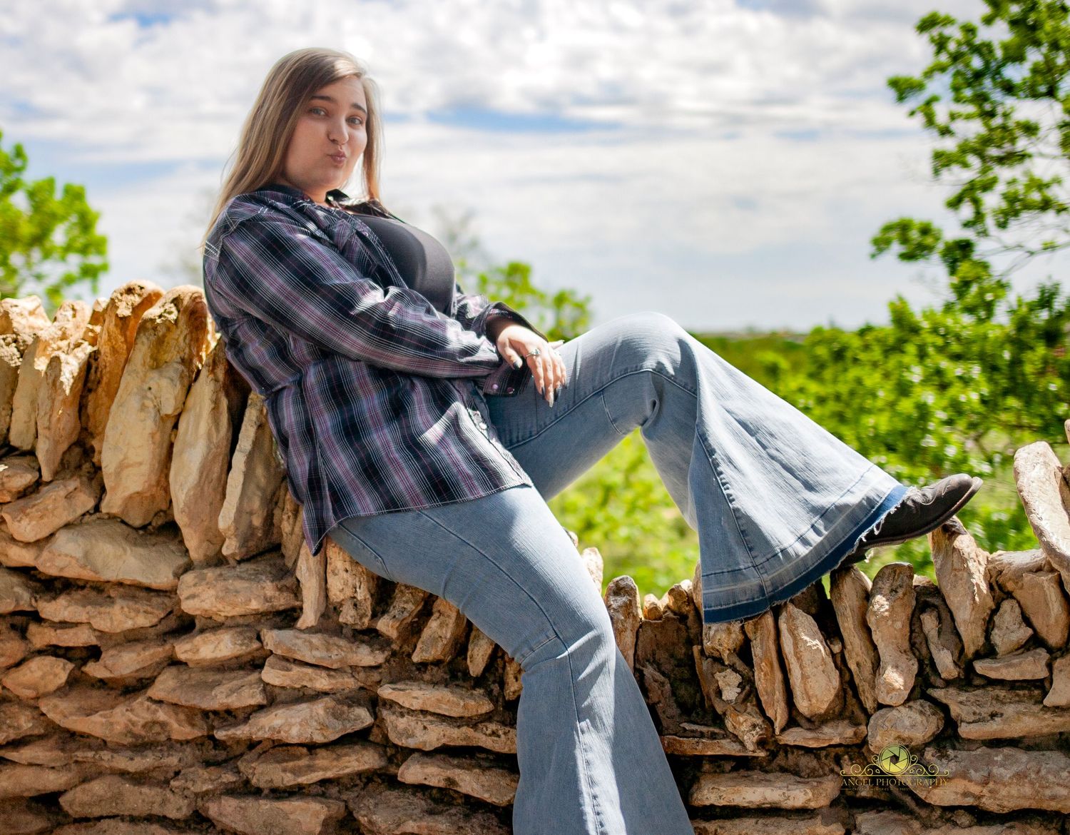 Senior portrait against stone wall wearing plaid shirt and flared jeans.
