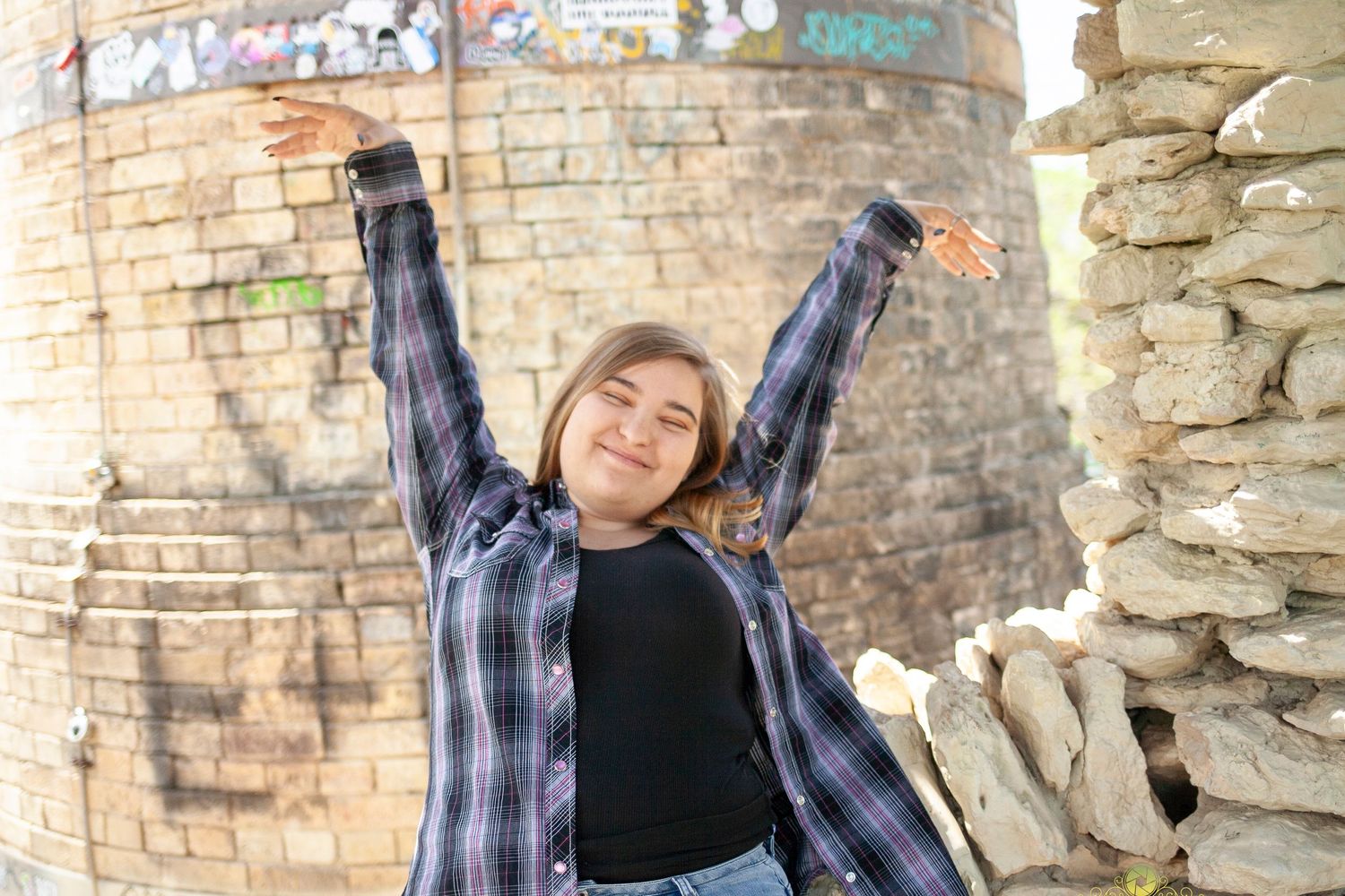 Person in plaid shirt poses playfully with arms raised against brick wall in outdoor setting.