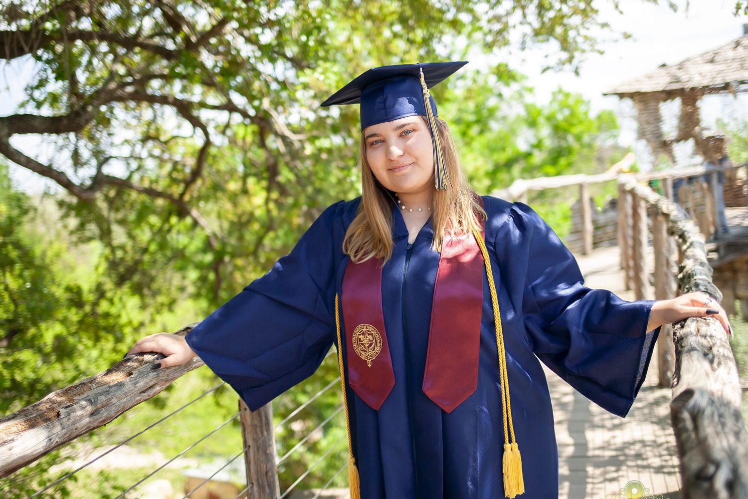Graduate in navy blue cap and gown with red stole makes peace sign outdoors.