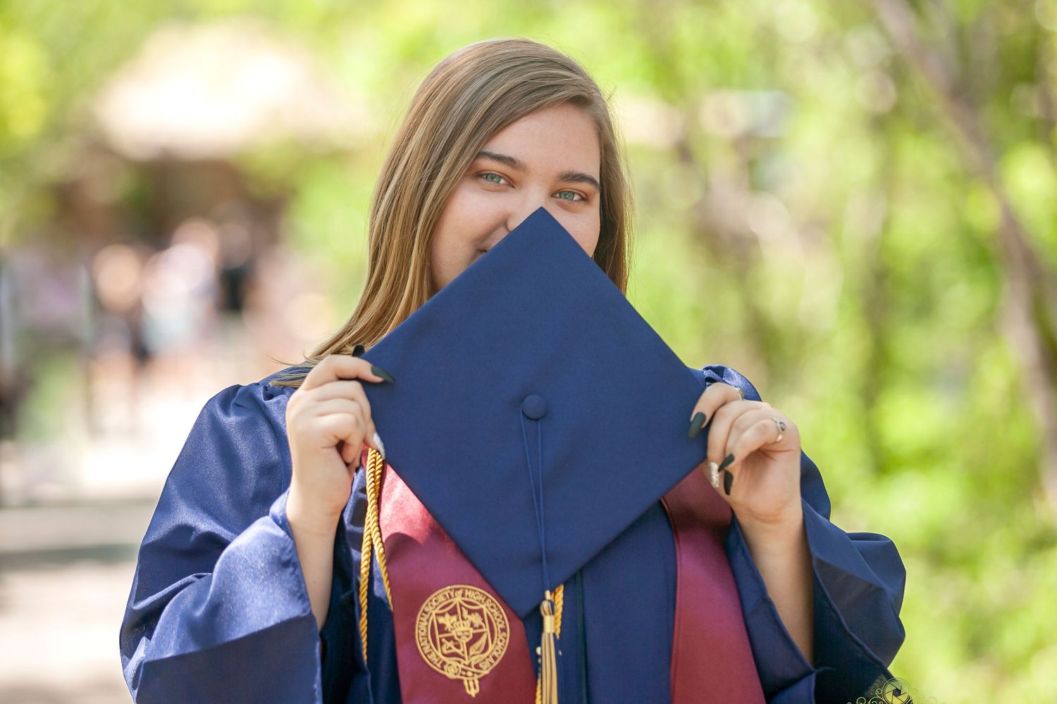 Graduate in blue regalia holds up mortarboard cap in front of face.