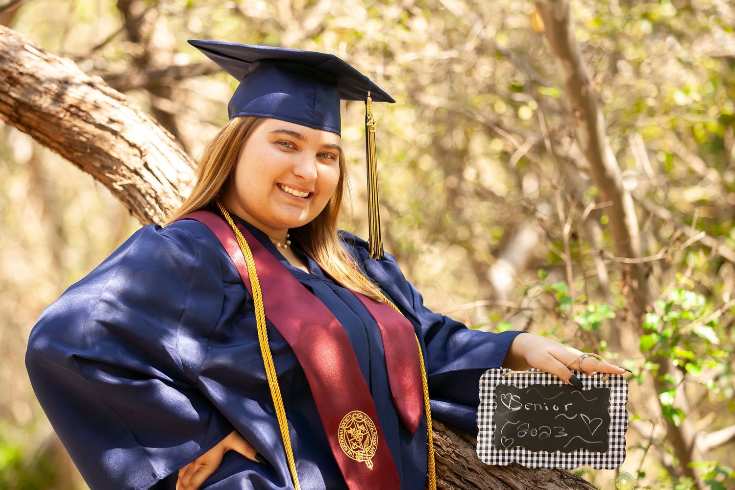 Graduate in academic regalia poses with diploma holder in outdoor wooded setting.