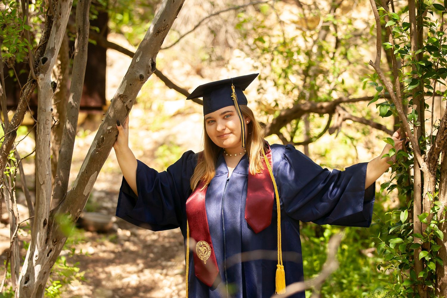 Graduate in blue cap and gown poses with outstretched arms among trees.