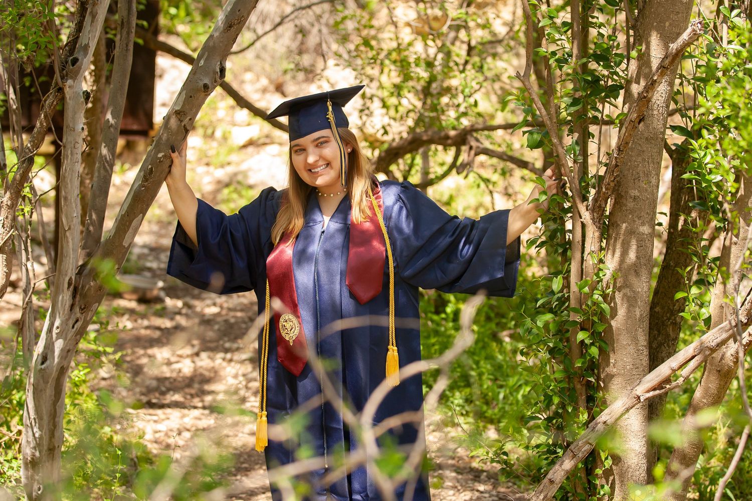Graduate in full regalia poses joyfully on wooded path during photo session.