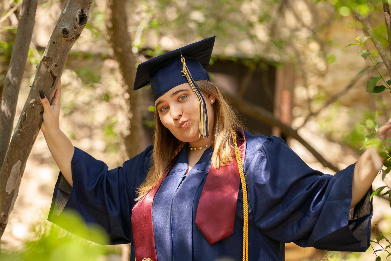 Graduate in academic dress poses expressively between tree branches.