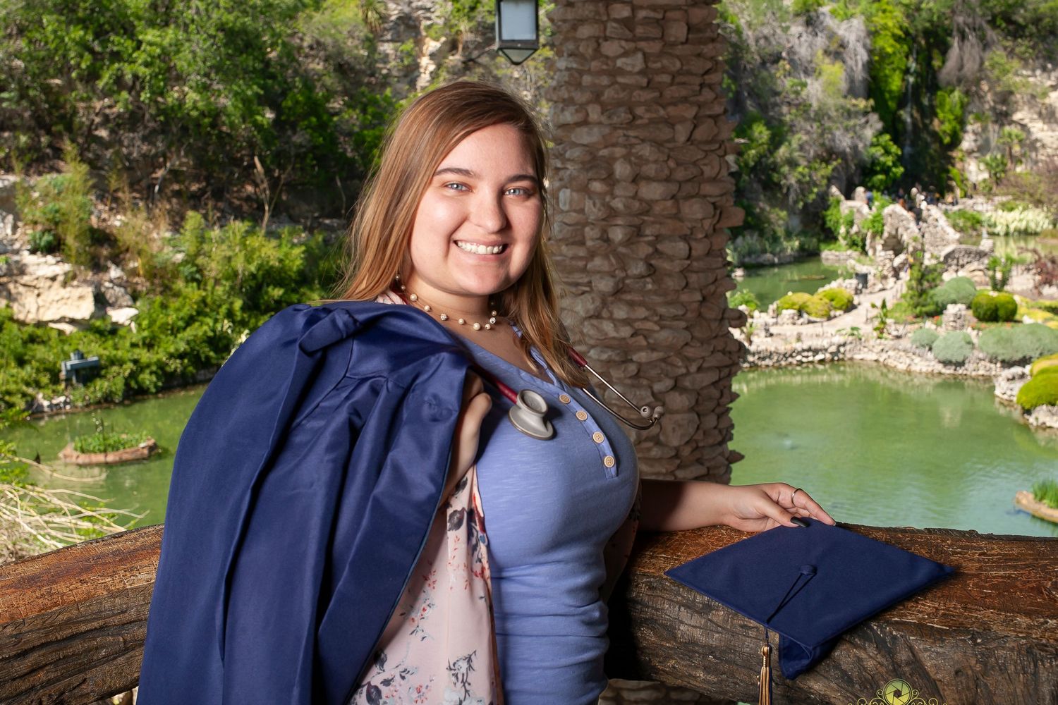A navy blue graduation gown against a scenic garden backdrop with a pond and stone features.