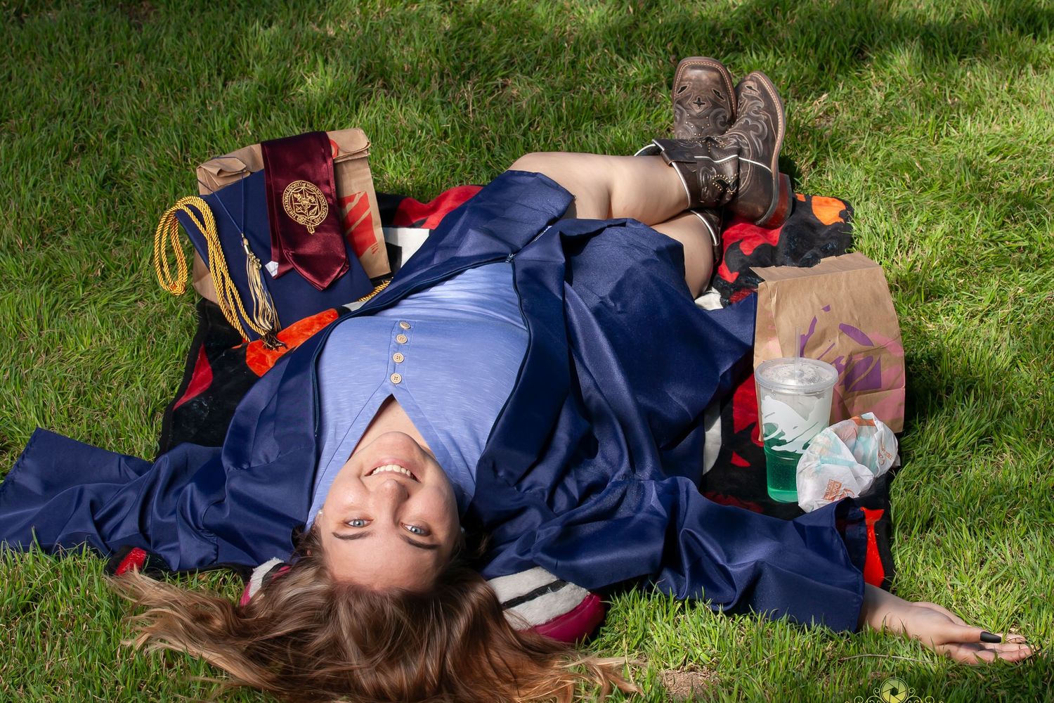 A graduate lying on grass with backpack and graduation attire.