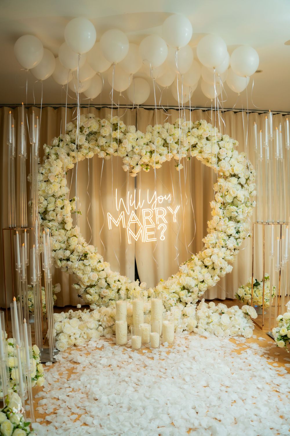 Romantic proposal setup with white balloons, floral arrangements, and neon signs in an elegantly decorated indoor space.
