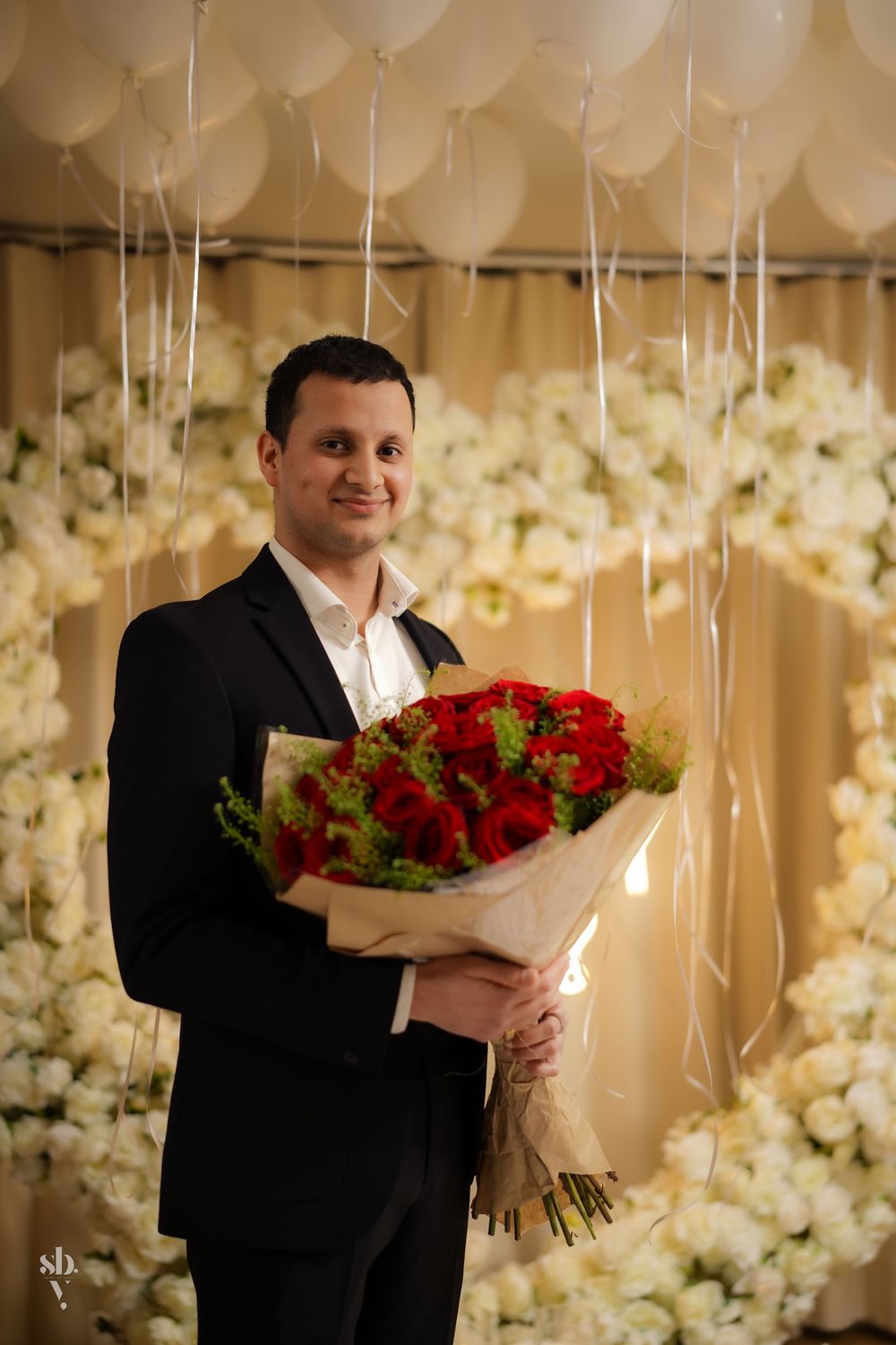 A black-suited person holding a vibrant bouquet of red roses in a room decorated with white balloons and floral arrangements.