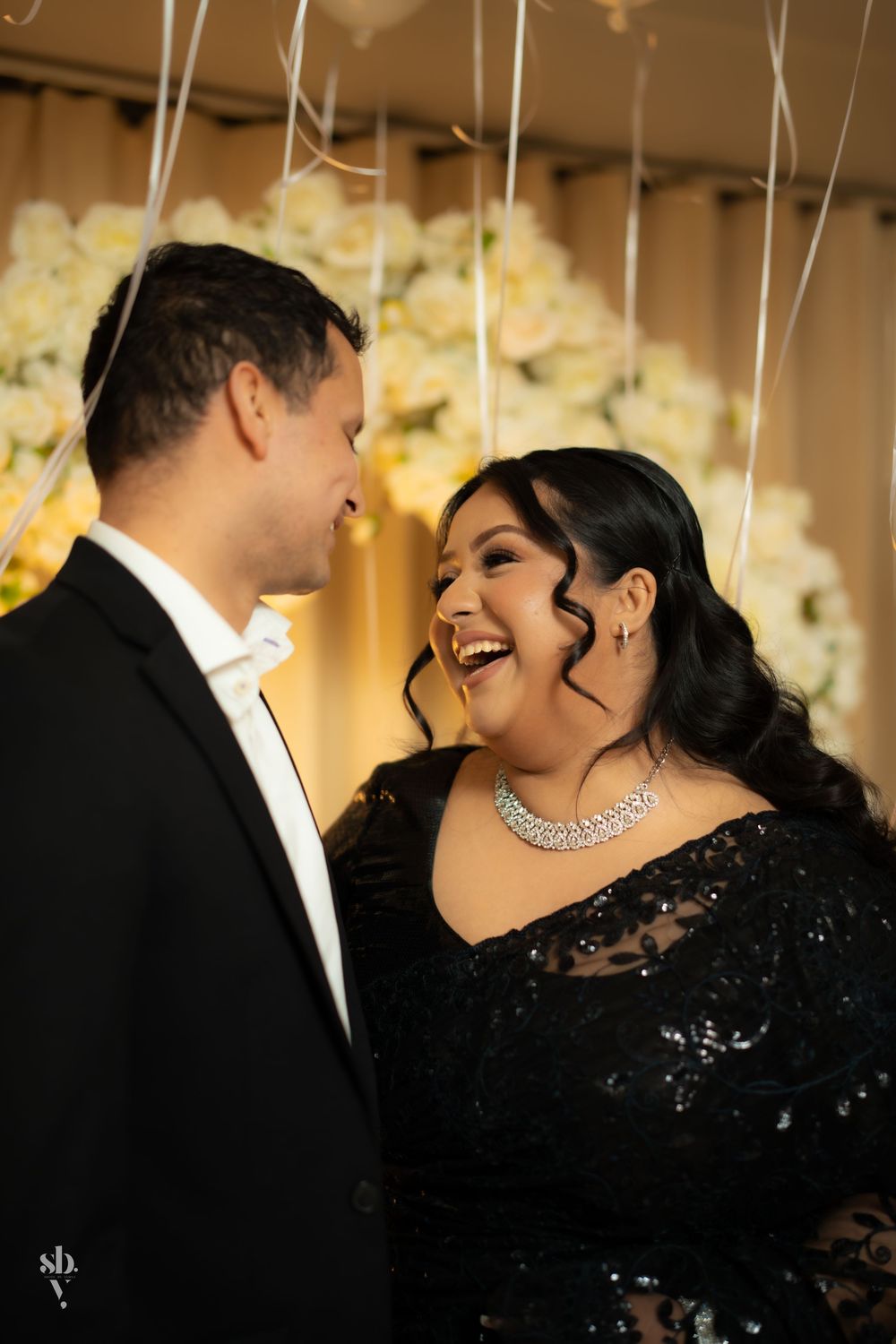 Formal couple share a joyful moment at an elegant event with white balloons and floral decorations in the background.