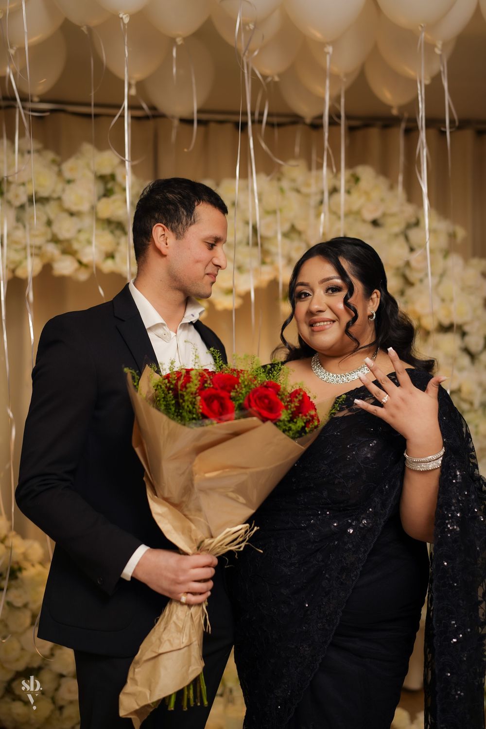 A couple shares a romantic moment with white balloons overhead and red roses as the backdrop features elegant floral decor.