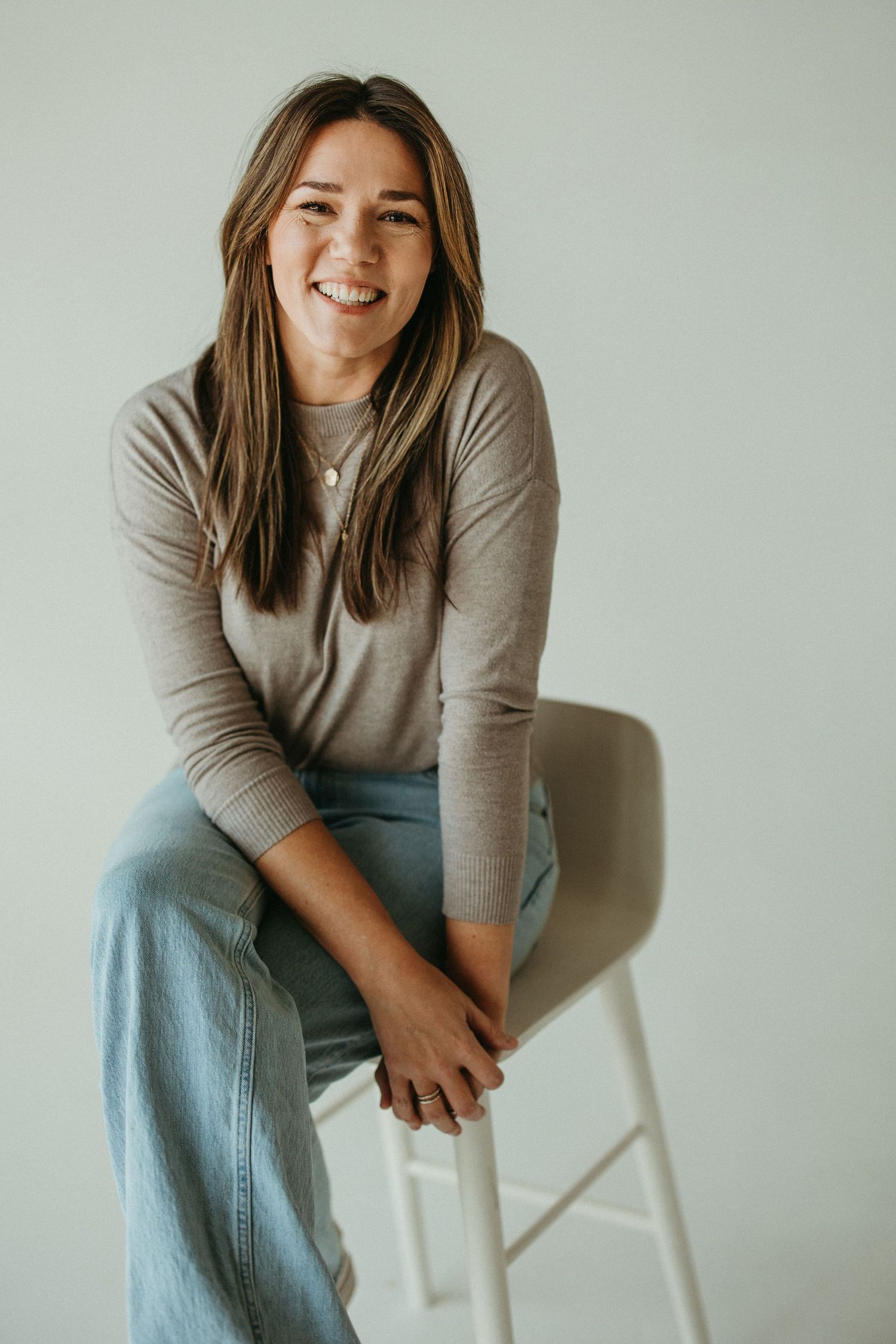 Portrait of a brunette woman smiling in a white studio space while sitting on a high stool.