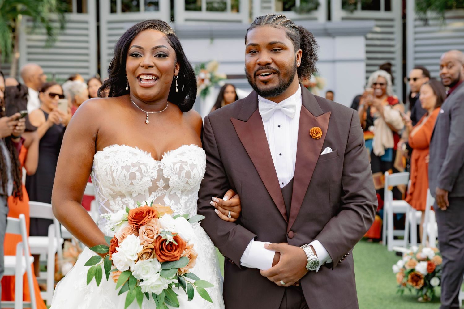A joyful bride and groom walk down the aisle at an outdoor wedding ceremony with guests applauding.