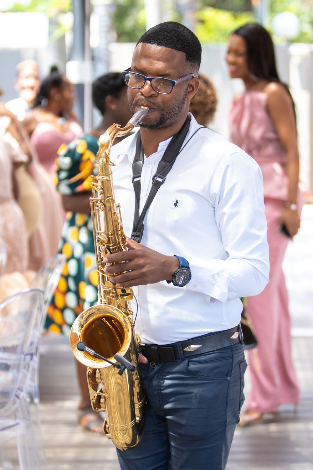 Musician playing saxophone while wearing white shirt and glasses at outdoor event.