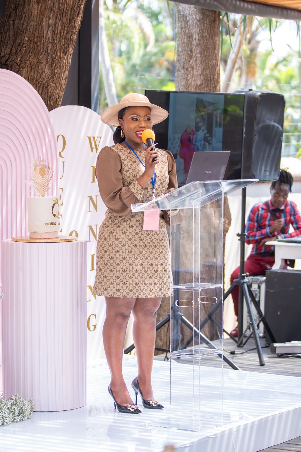 Speaker at podium wearing tan dress and hat during outdoor presentation.