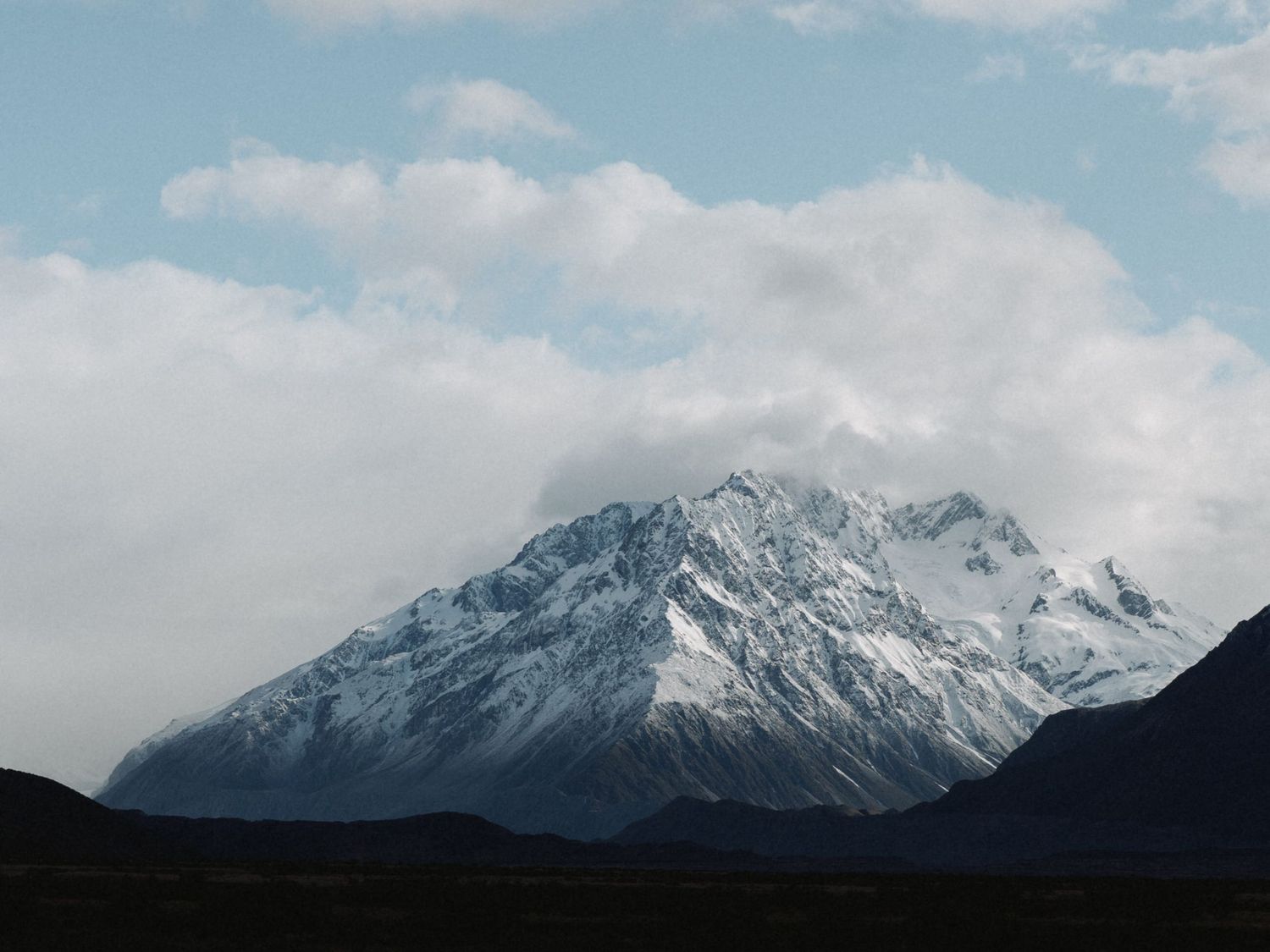 A majestic snow-covered mountain peak rises against a cloudy blue sky.