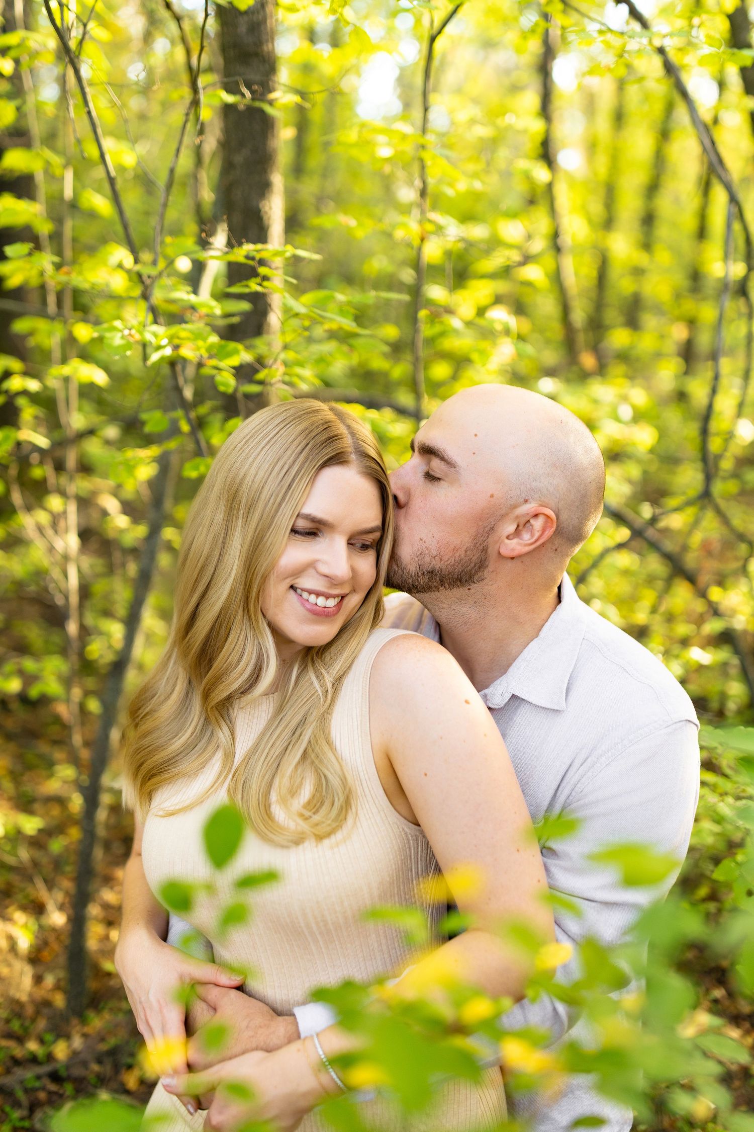 Intimate couple moment captured in sunlit forest with green foliage.
