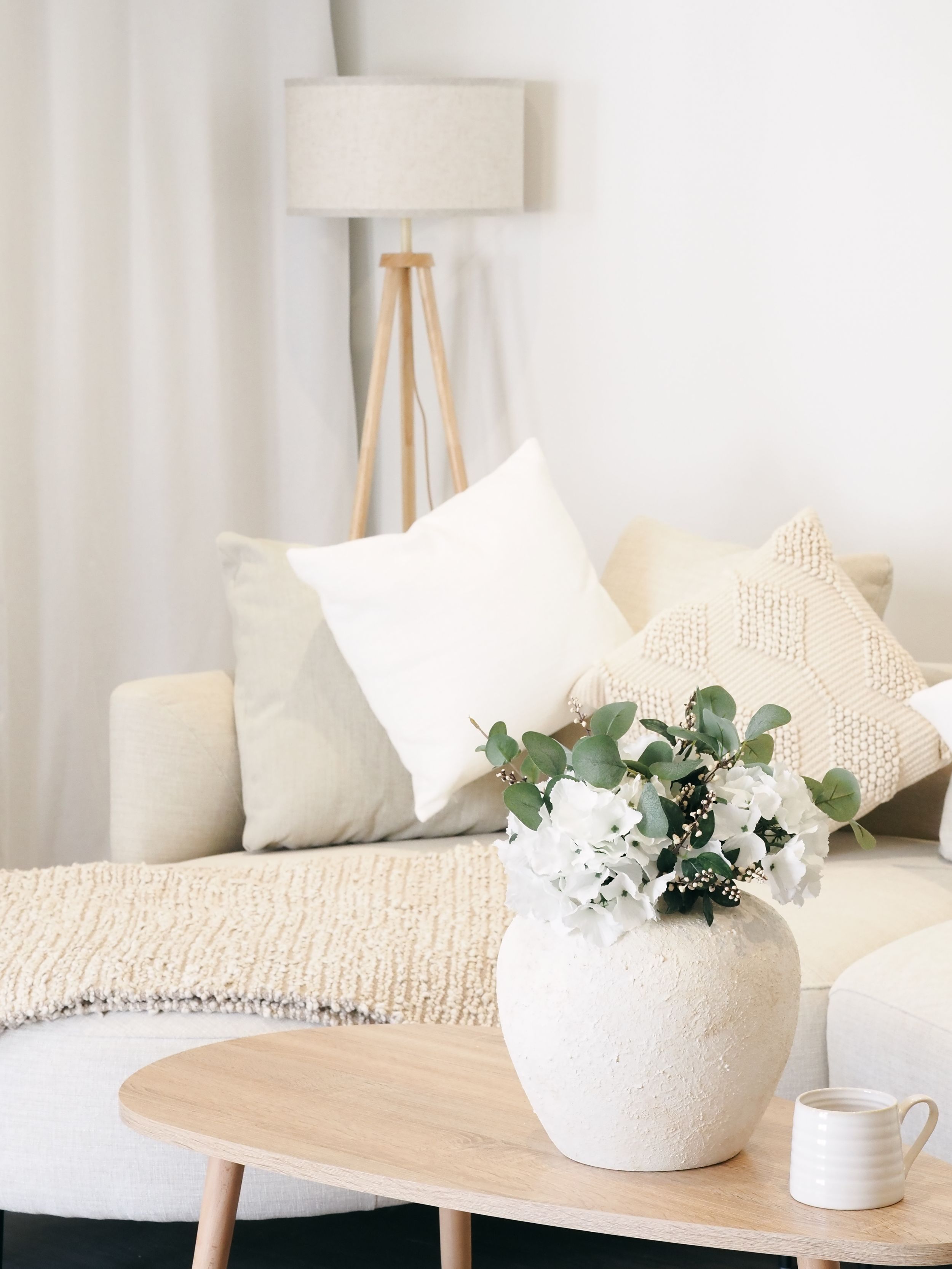 Cozy living room corner with tripod floor lamp, beige throw pillows, and white floral arrangement on wooden coffee table.