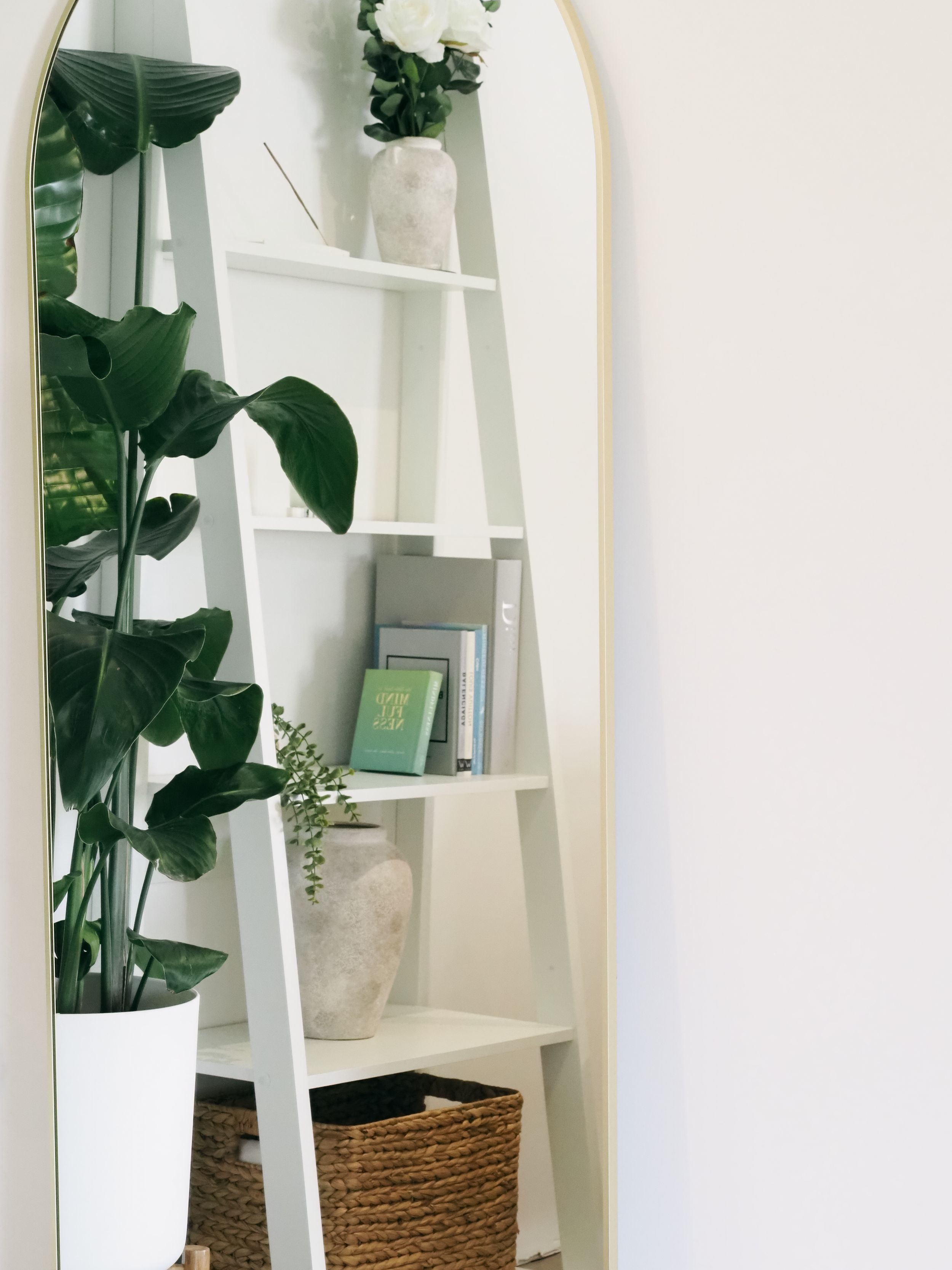 White ladder shelf with potted plants, woven basket, and books arranged against a light wall with arched mirror.