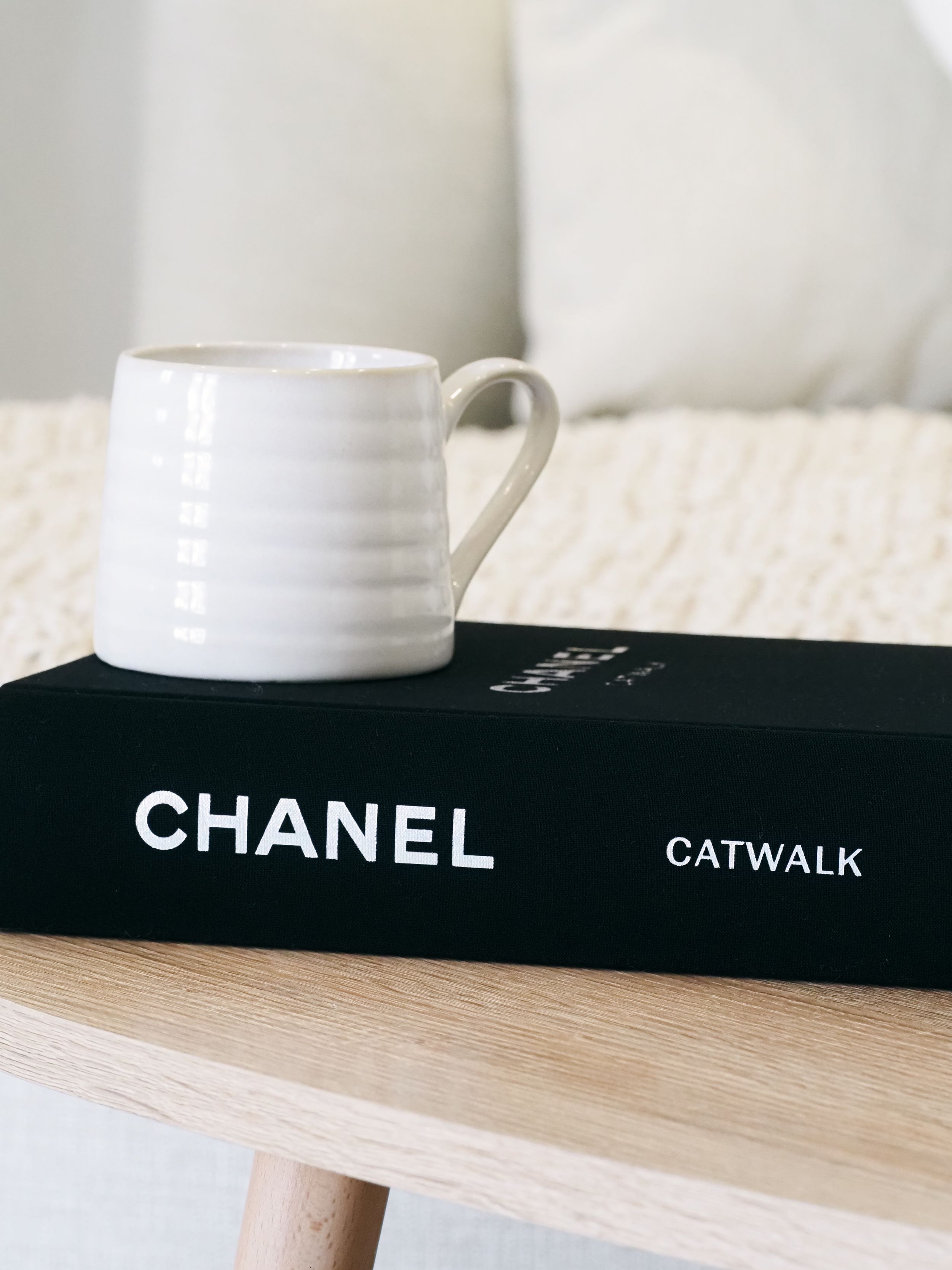 A white ceramic coffee mug sits on a black Chanel Catwalk fashion book on a light wooden surface.