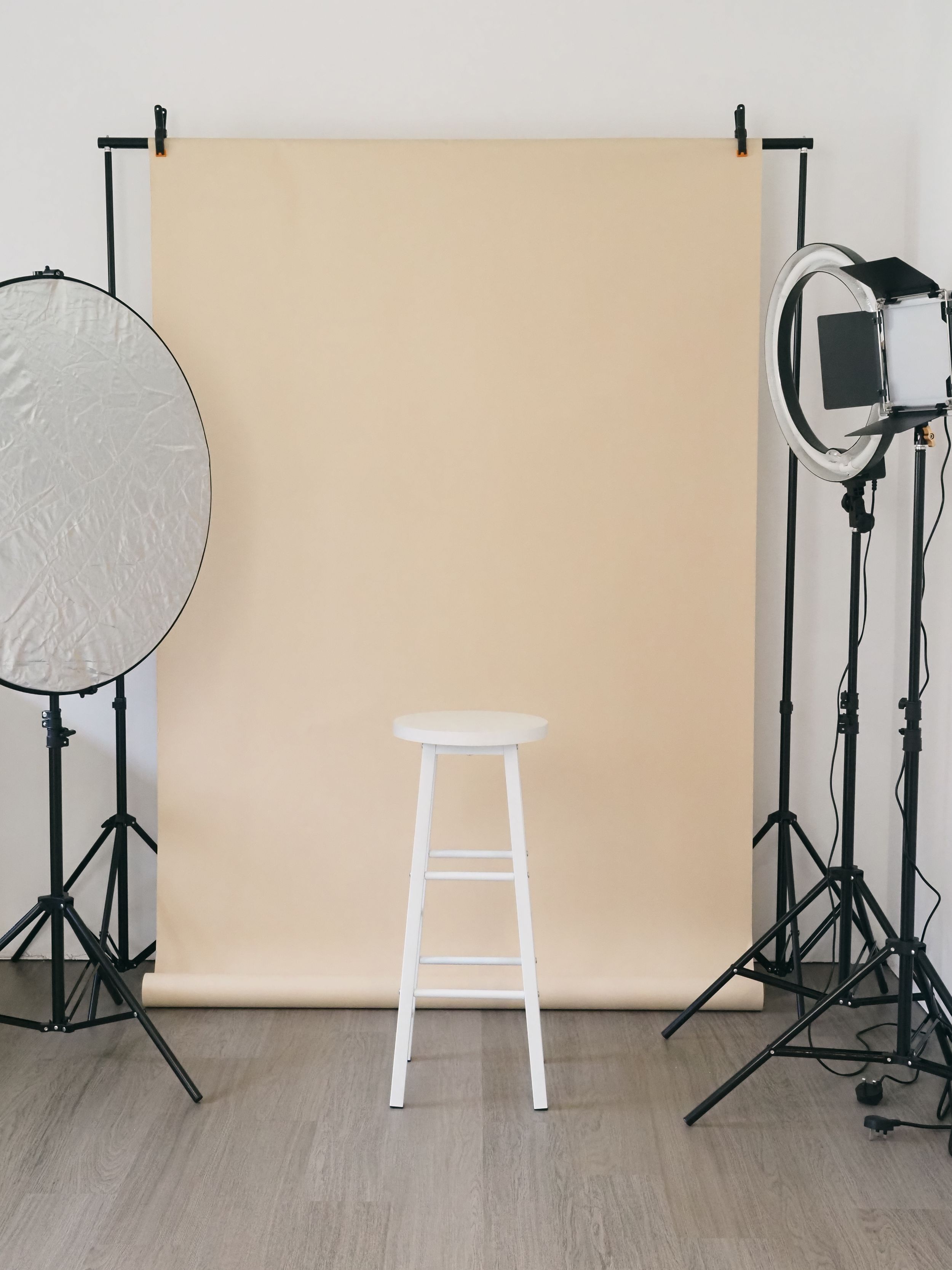 Photography studio setup with lighting equipment, reflector, cream backdrop, and white wooden stool against a hardwood floor.
