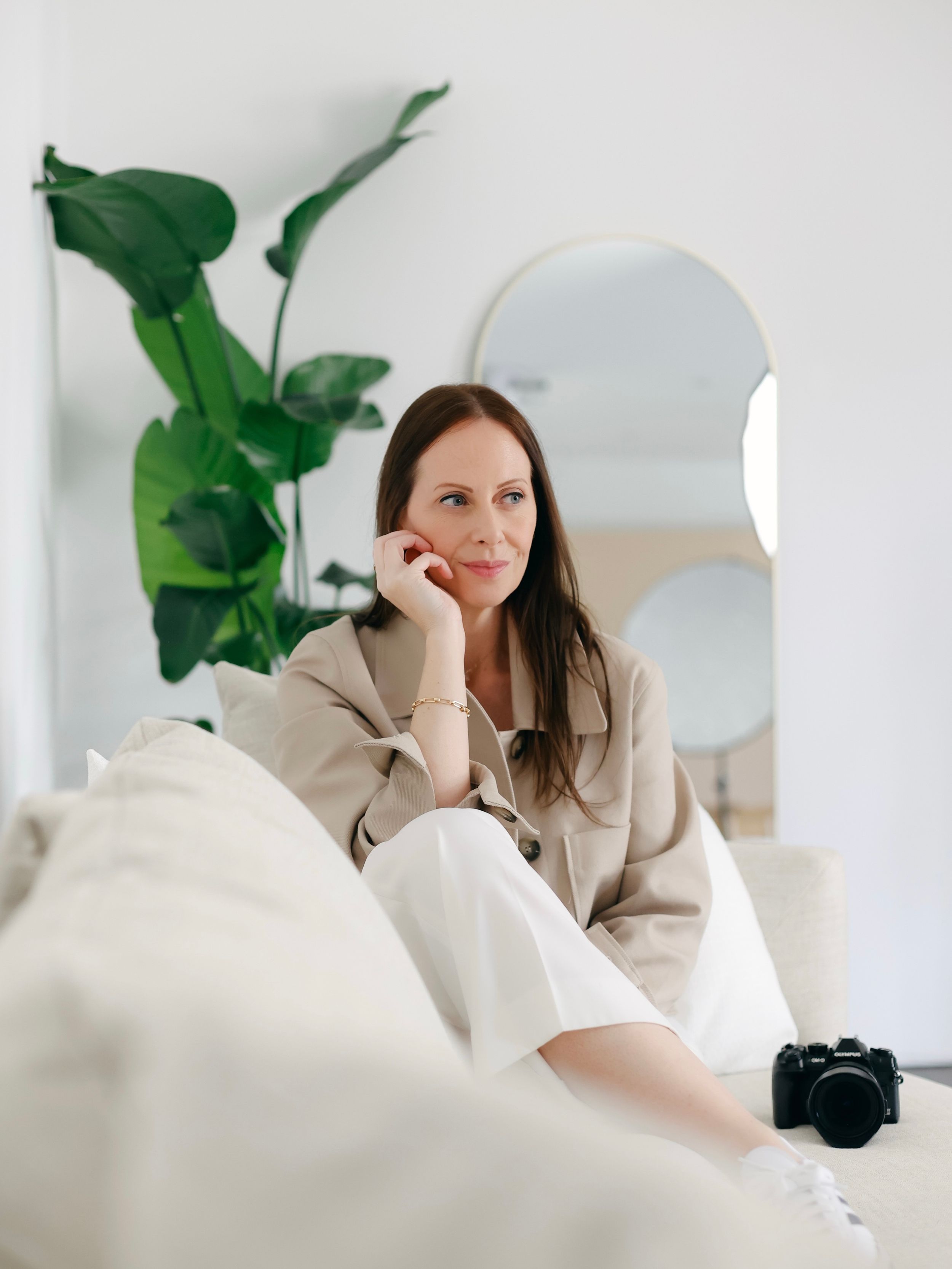 Relaxed figure lounges on white couch with a camera nearby, next to large houseplant and arched mirror in minimalist setting.