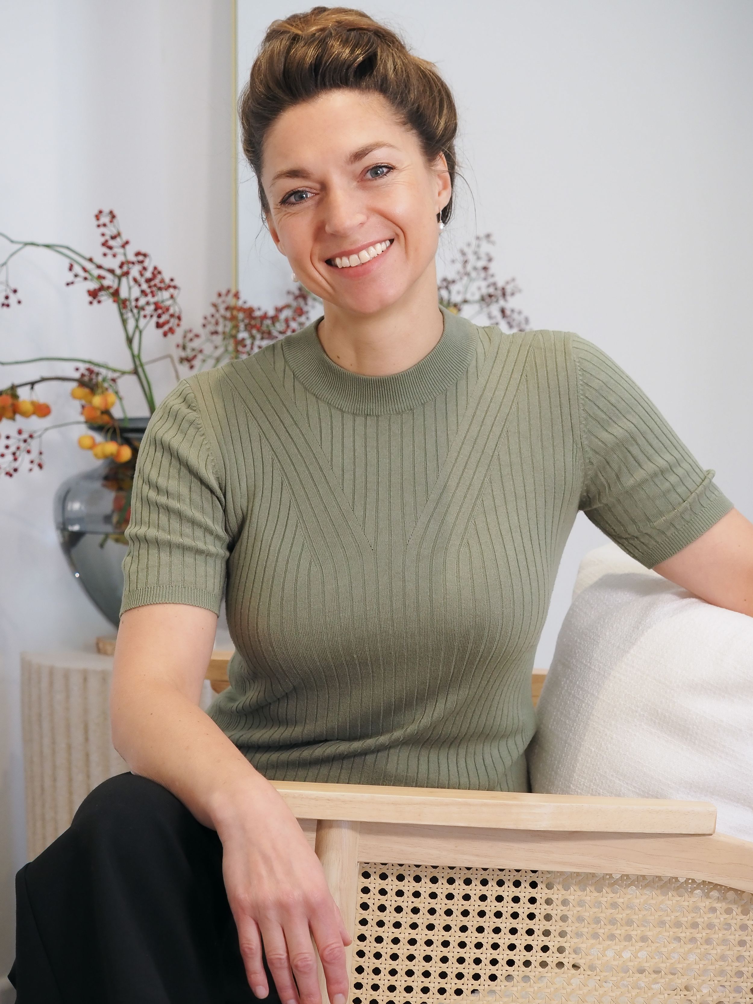 Smiling woman in sage green ribbed knit top seated in cane chair with floral decor in bright modern interior.