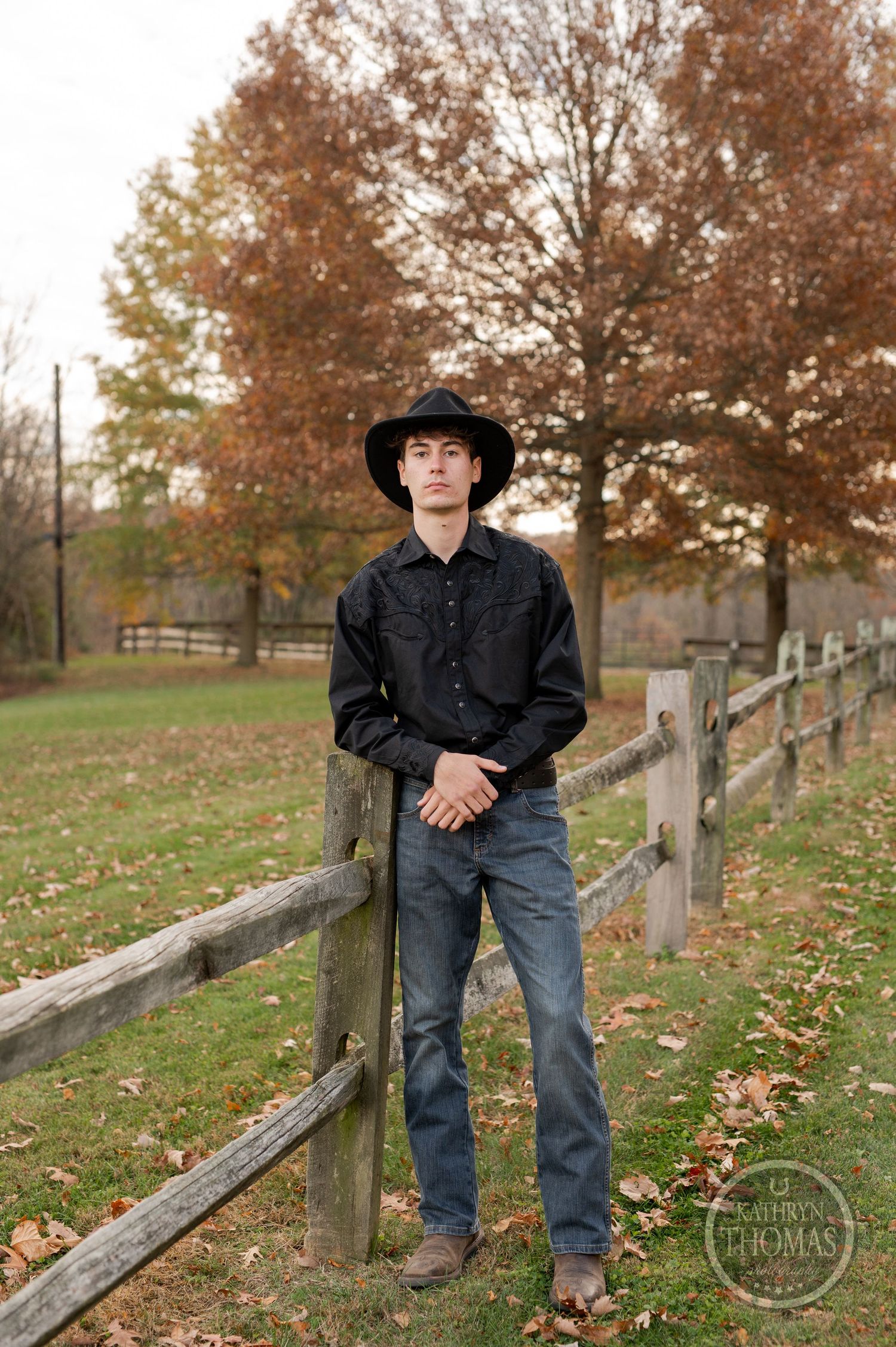 A western-styled portrait against a rustic wooden fence with autumn trees in the background on a cloudy day.