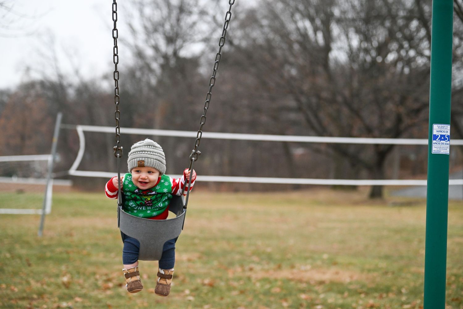 A photograph of toddler Nolan sitting in a swing at the park, bundled up and laughing while smiling big at the camera