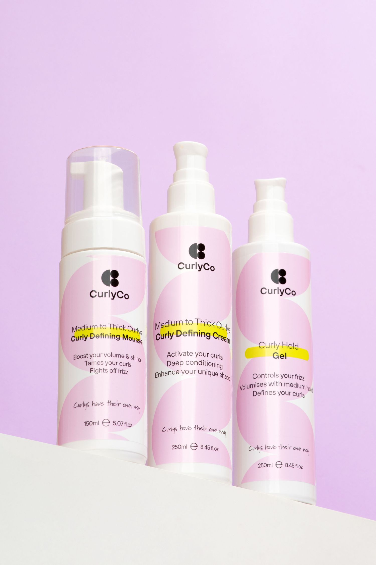 Three pink CurlyGo hair styling products lined up  as a hero shot against pastel purple background. Shot from a low angle looking up.