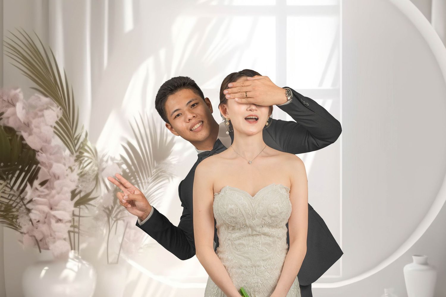 Playful wedding portrait with couple in formal attire against white circular backdrop with palm leaves.
