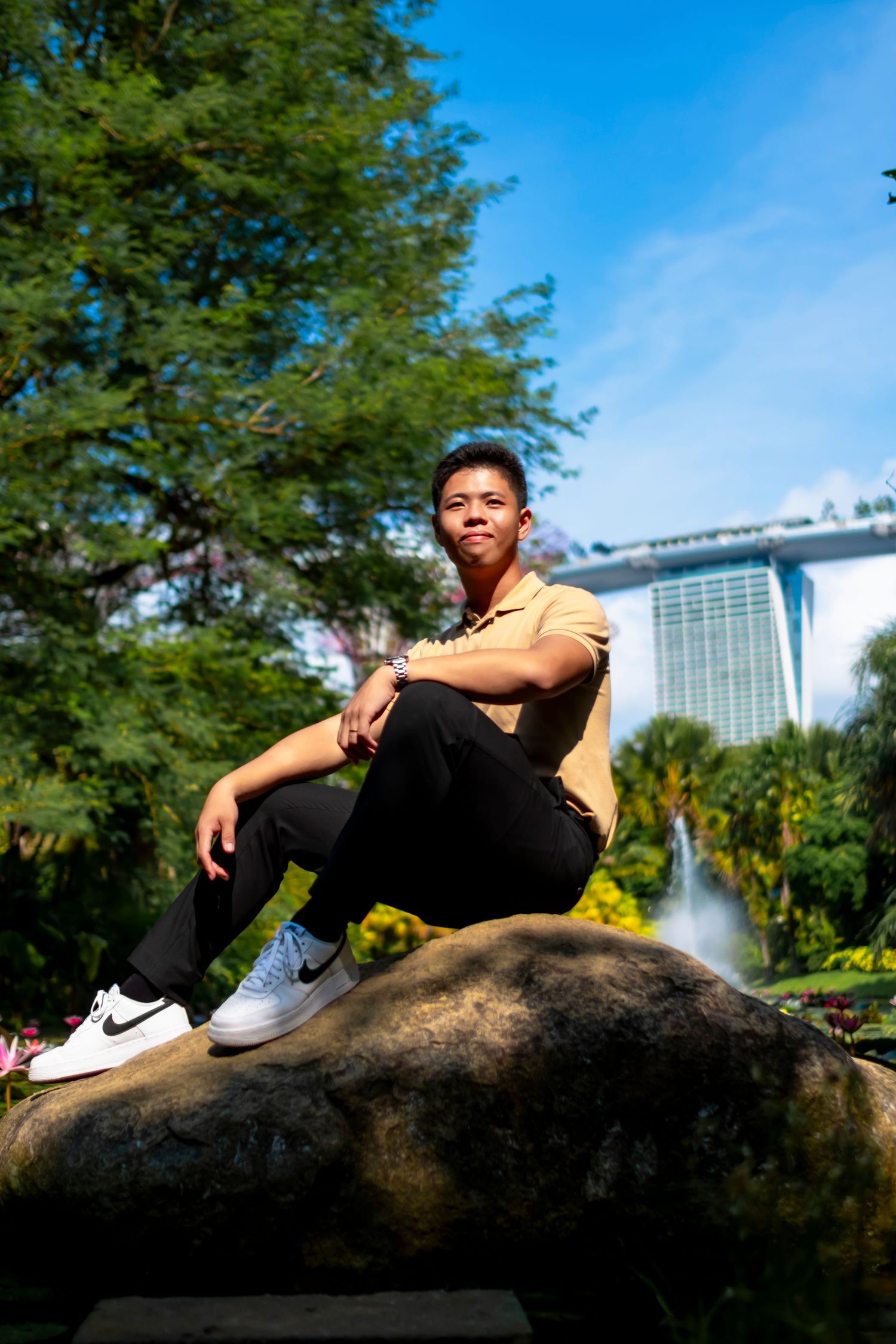 Person in beige polo shirt sitting on rock with urban backdrop and blue sky.