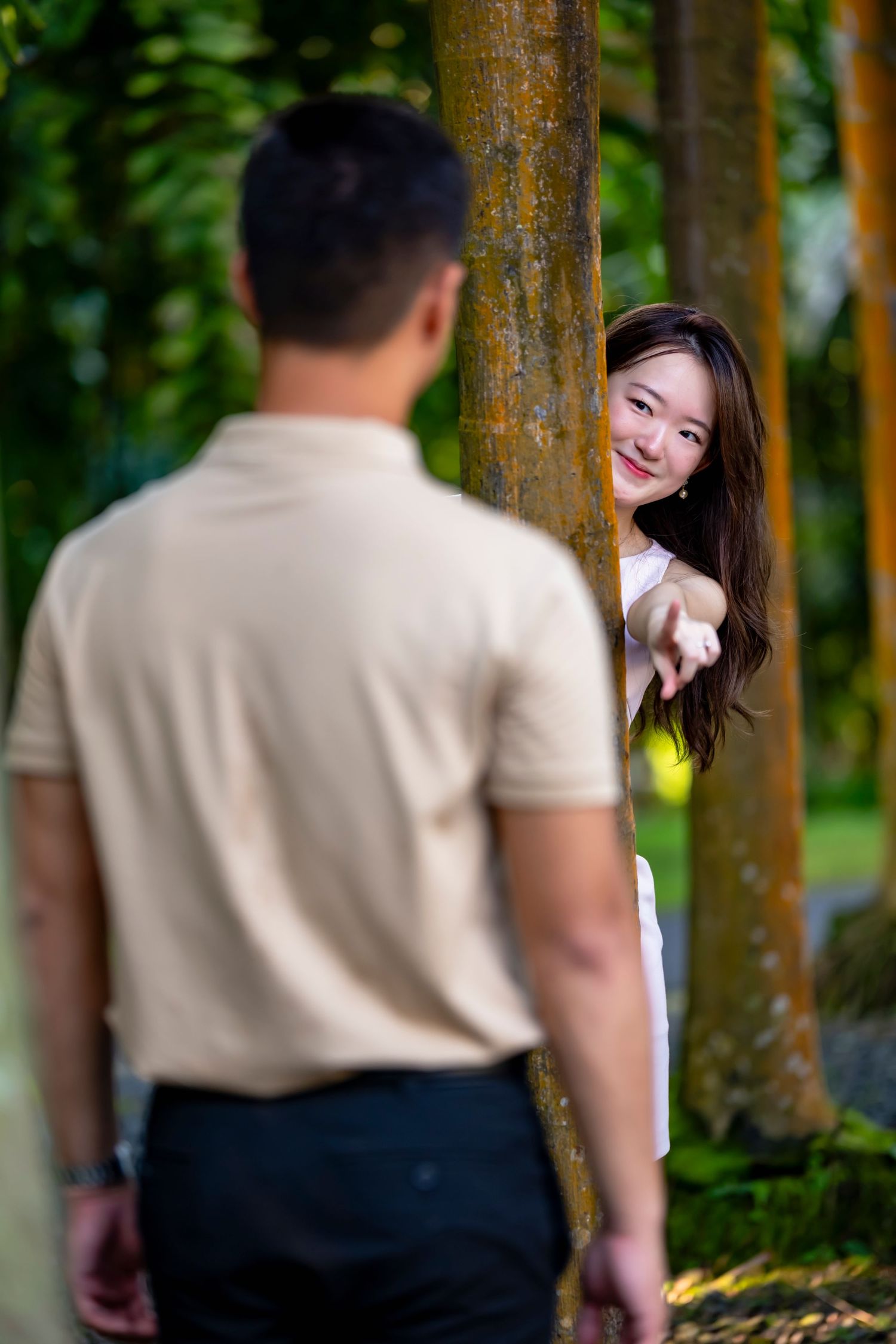 Woman peeks playfully from behind tree trunk in outdoor garden setting.