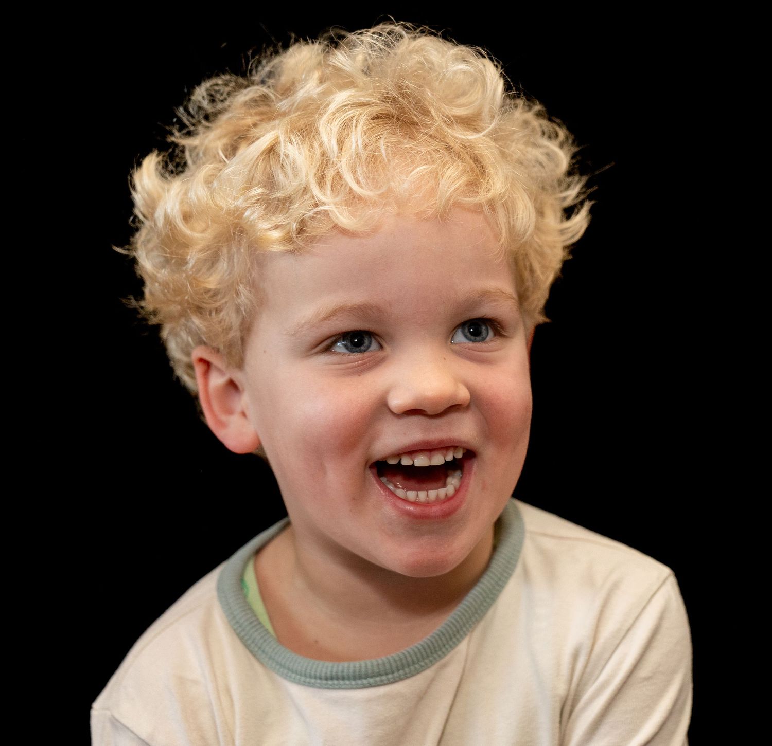 "Blonde peuter met wilde krullen lacht vrolijk op een zwarte achtergrond. Zijn gezicht straalt plezier uit."