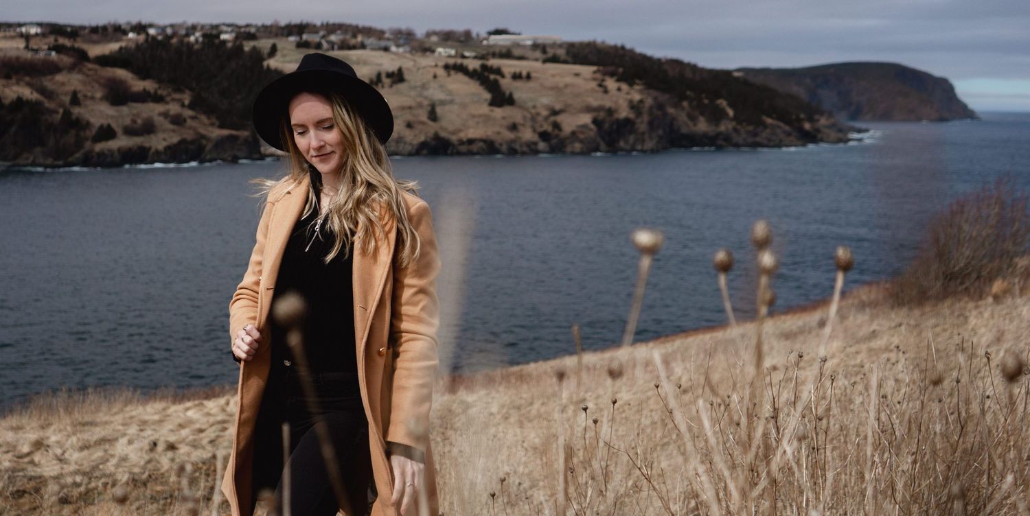 Person in tan coat and black hat stands along coastal cliffside with scenic ocean bay and rolling hills in background.