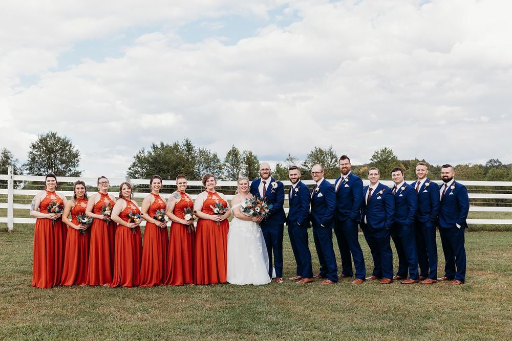 full wedding party with bridesmaids in orange dresses, bride and groom, groomsman in blue suits during outdoor photos