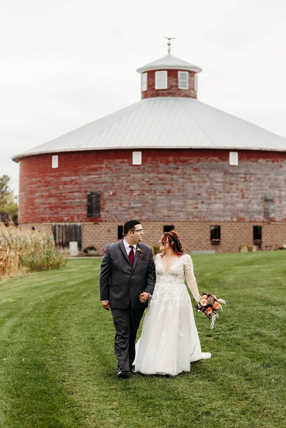 bride and groom casually walking towards camera with round barn in the backgrounds