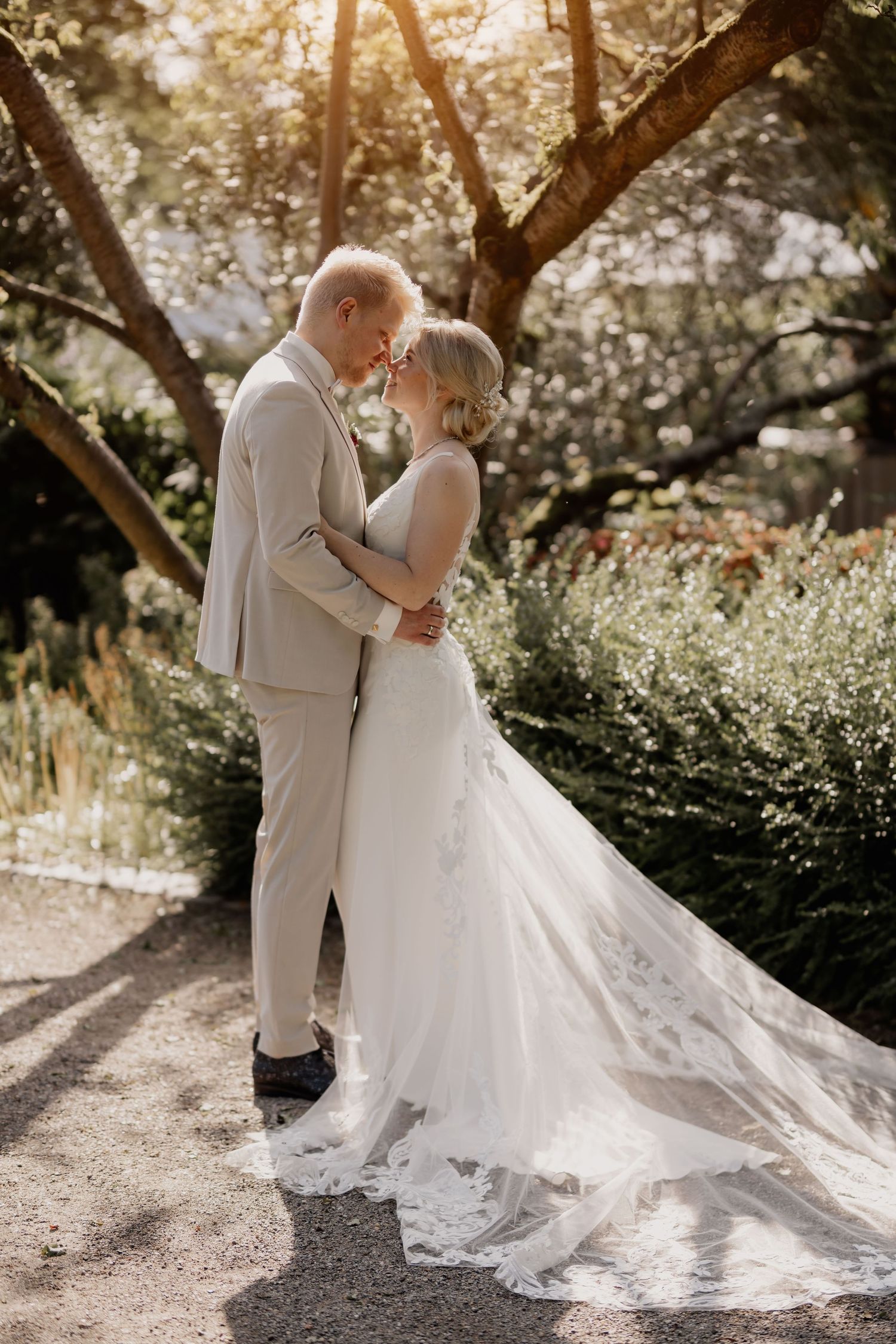 Romantic golden hour wedding portrait with couple in light colored attire against sunlit garden.