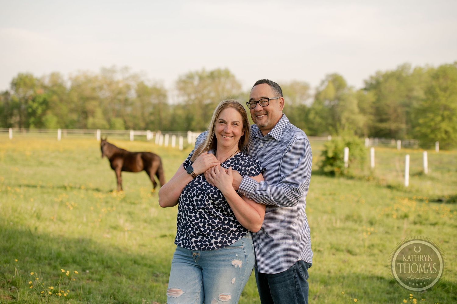 A couple embraces in a sunny field with a horse grazing in the background near white fence posts.