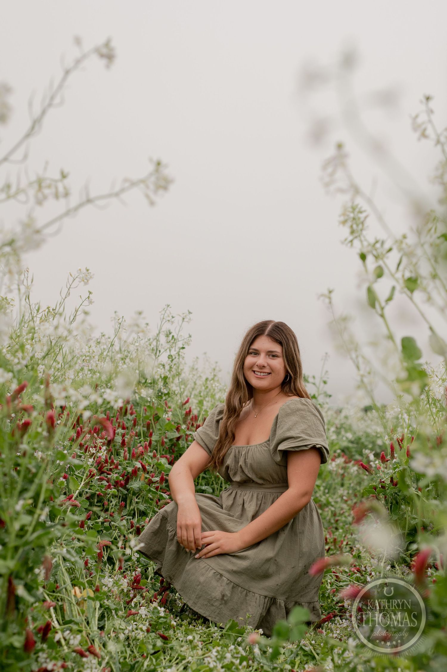 Someone in a sage dress sits contentedly among red and white wildflowers in a misty field.