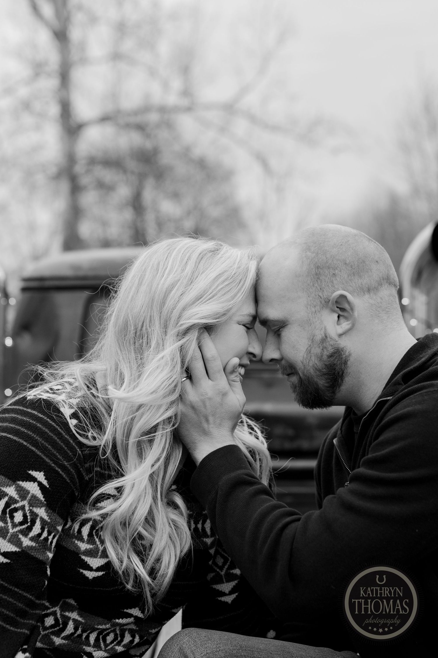 An intimate black and white portrait of a couple sharing a tender moment against a vintage truck backdrop.