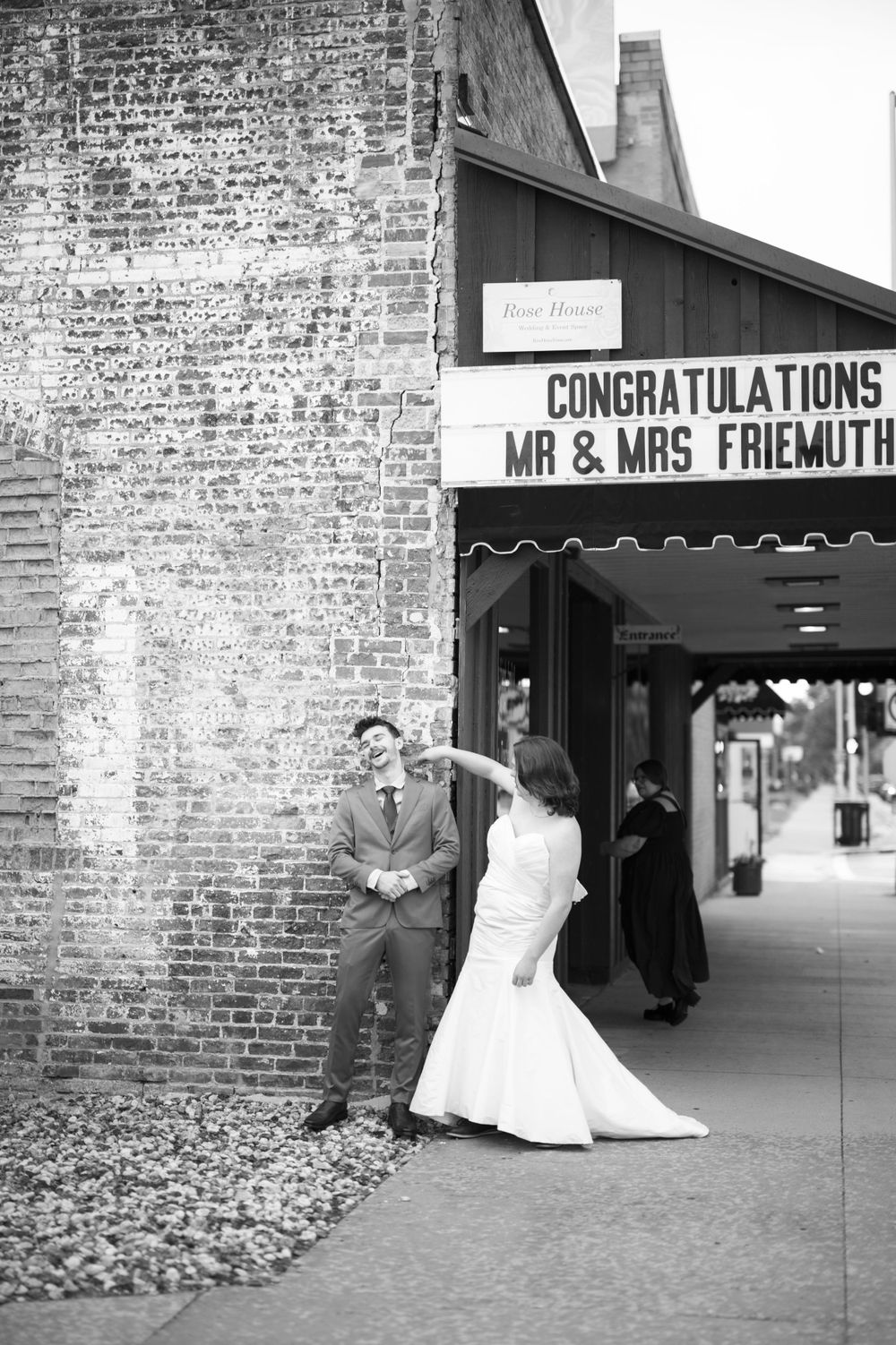 Black and white photo of a newlywed couple standing by a brick wall under a theater marquee with congratulatory message.