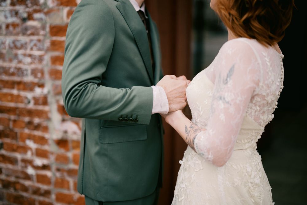 A couple holds hands during their wedding ceremony against a rustic brick wall background.