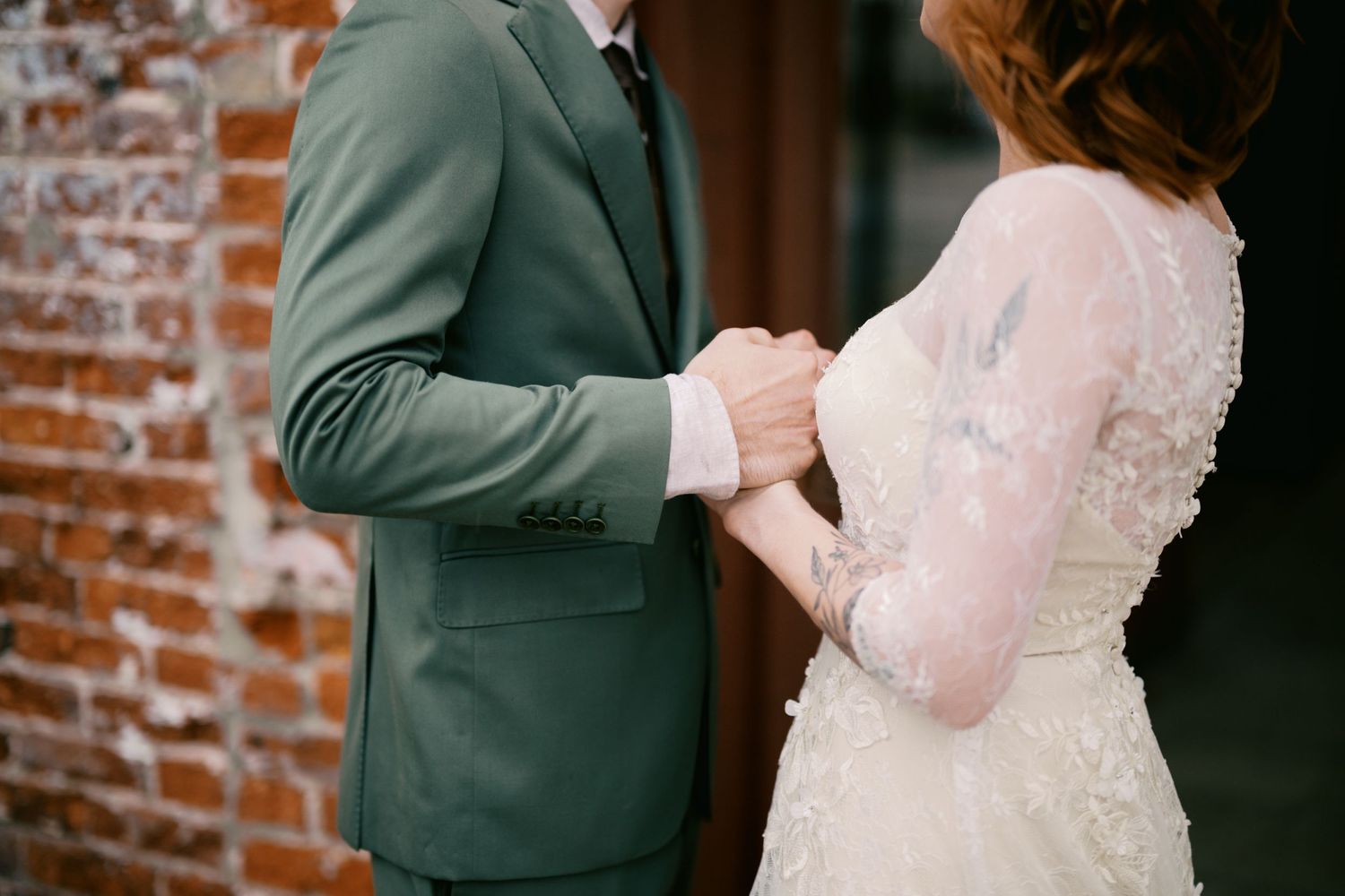 A couple holds hands during their wedding ceremony against a rustic brick wall background.