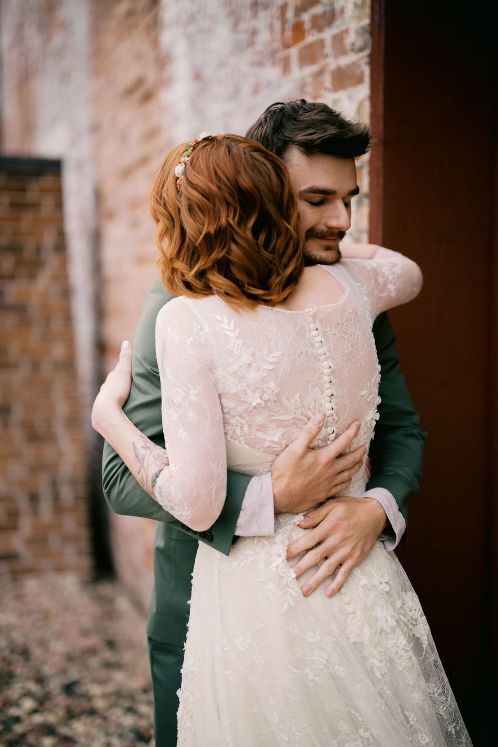 A wedding couple embraces tenderly against a rustic brick wall backdrop, the bride wearing a long-sleeved lace gown.