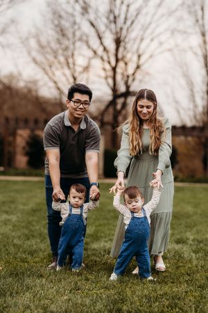 Parents helping twin toddlers walk on green grass in a park setting.