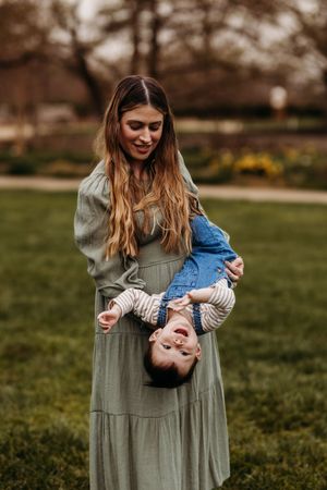 Smiling mother in green dress playfully holding laughing baby upside down outdoors.