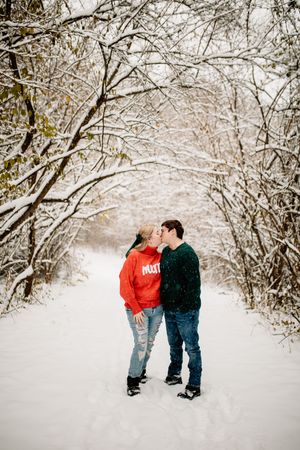 Couple sharing a kiss on a snow-covered forest trail, dressed in festive red and green sweaters.
