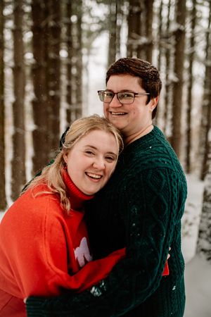 Happy couple embracing in snowy pine forest, woman in red sweater and man in green cable-knit.